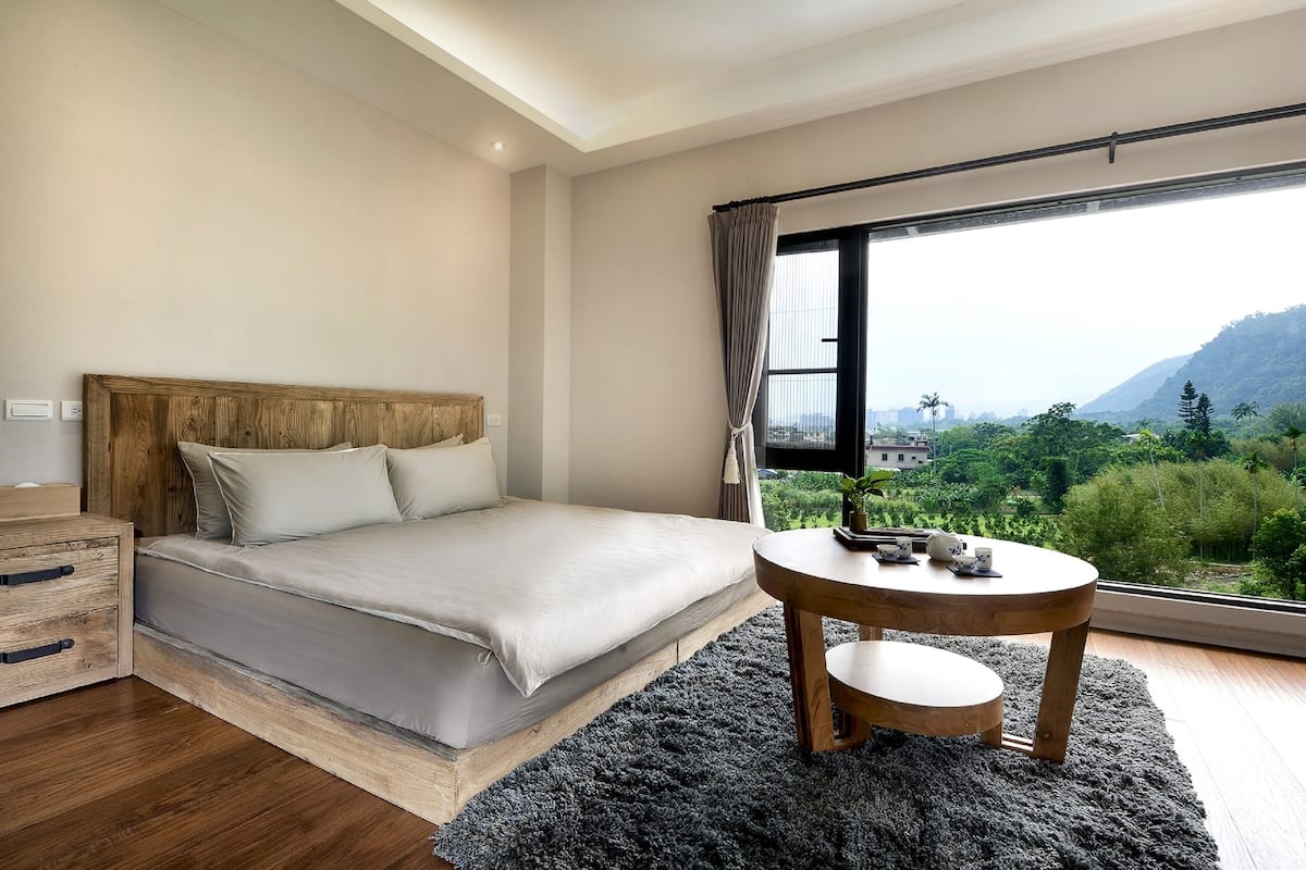 Ein Schlafzimmer mit einem großen Bett, einem hölzernen Nachttisch, einem runden Tisch und Blick auf Grünflächen und Berge durch ein großes Fenster.