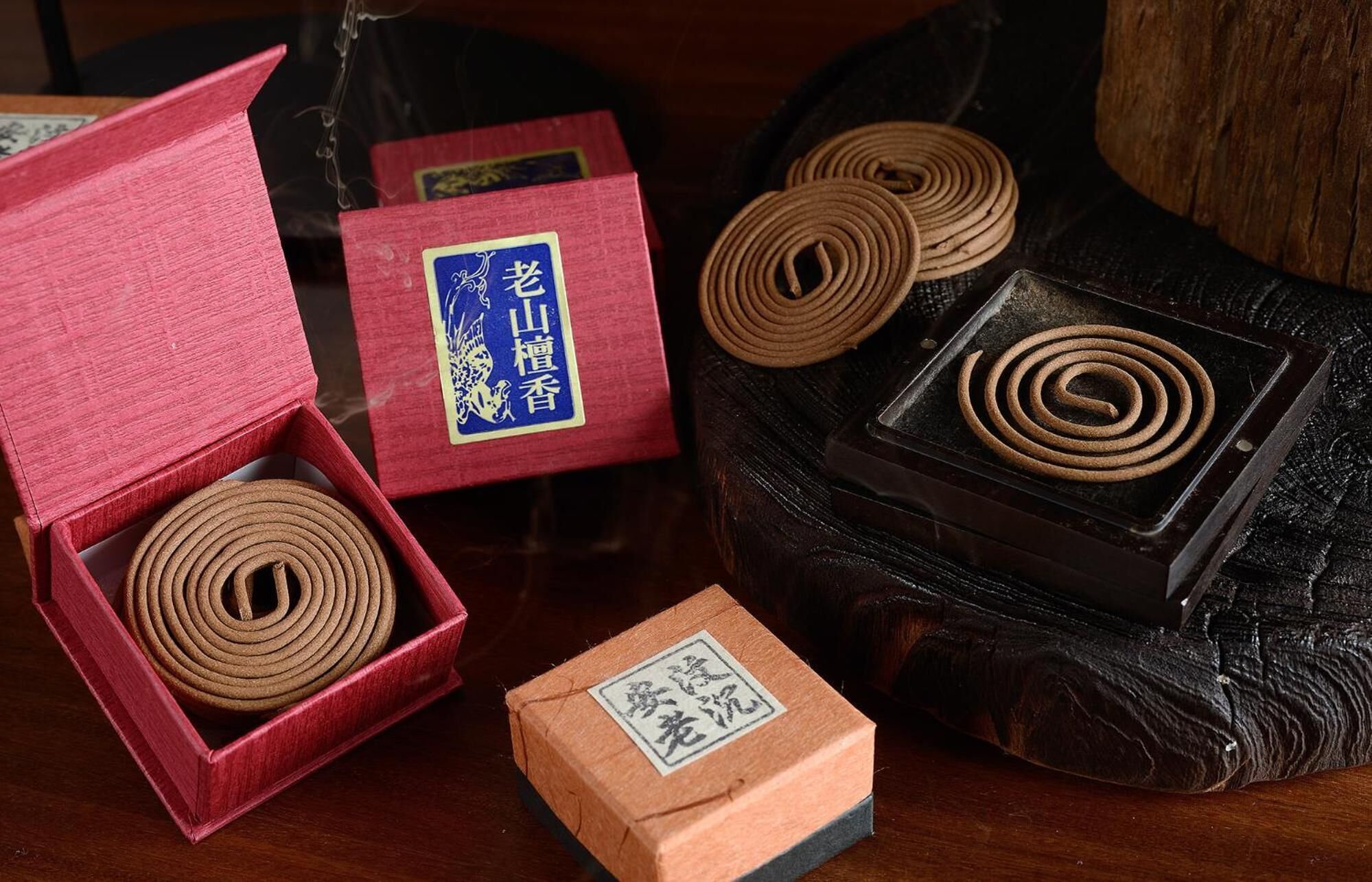 A wooden box with a red exterior and a blue label, a smaller wooden box with a label, and several coiled incense sticks on a dark surface.