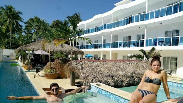 A swimming pool area with a thatched-roof structure, palm trees, and a multi-story building with balconies.