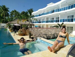 A swimming pool area with a thatched-roof structure, palm trees, and a multi-story building with balconies.