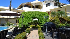 A poolside area with lounge chairs, a covered grill, and a pathway leading to a building with a thatched roof.