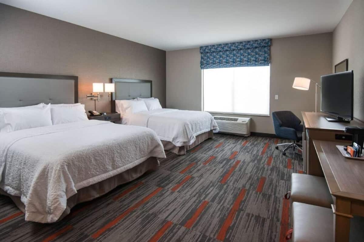 A hotel room with two beds, a desk, a chair, a TV, a lamp, and a window with a blue valance.