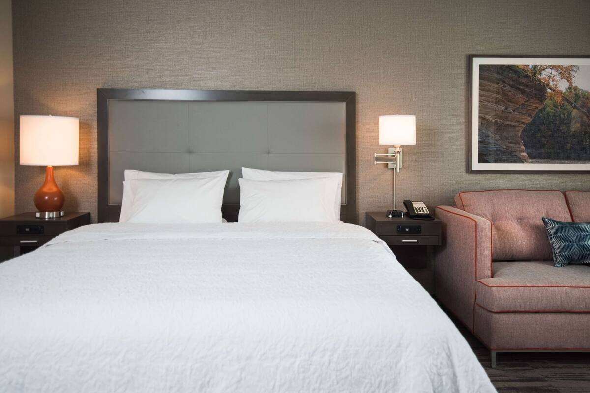 A hotel room with a large bed, two bedside tables with lamps, a nightstand with a phone, a sofa with a pillow, and a framed picture on the wall.