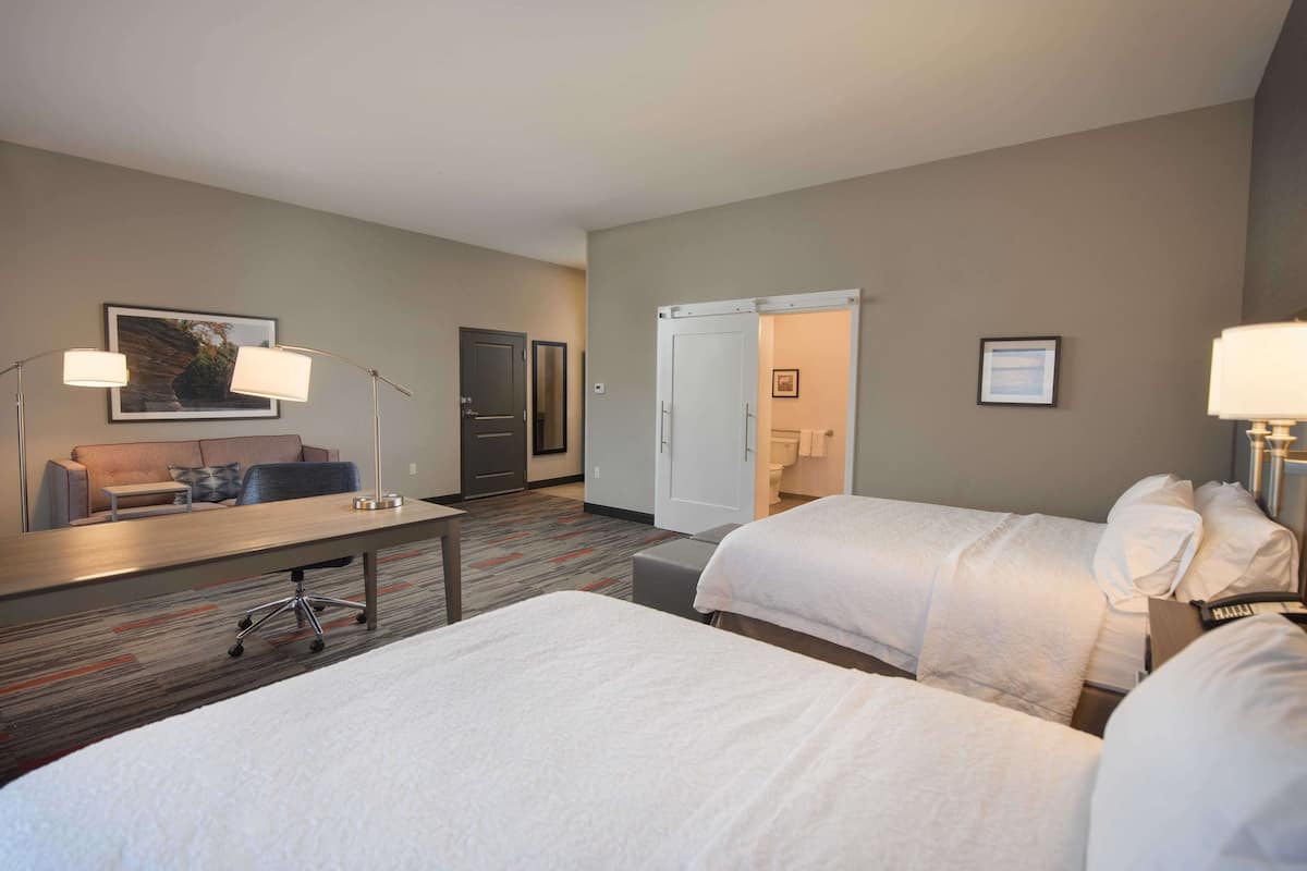 A hotel room with two beds, a desk with a chair, a sofa, and a door leading to a bathroom.