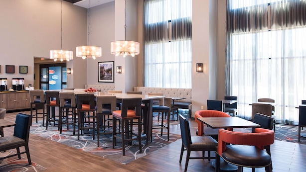 A hotel lobby with a central seating area, multiple tables, and chairs. There are large windows with sheer curtains, a ceiling fan, and a coffee station. - Hampton Inn & Suites at Wisconsin Dells Lake Delton (Baraboo)