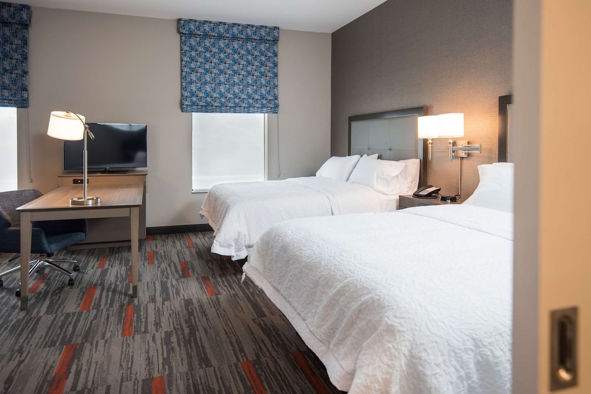 A hotel room with two beds, a desk with a lamp, a television, and a window with a patterned curtain.