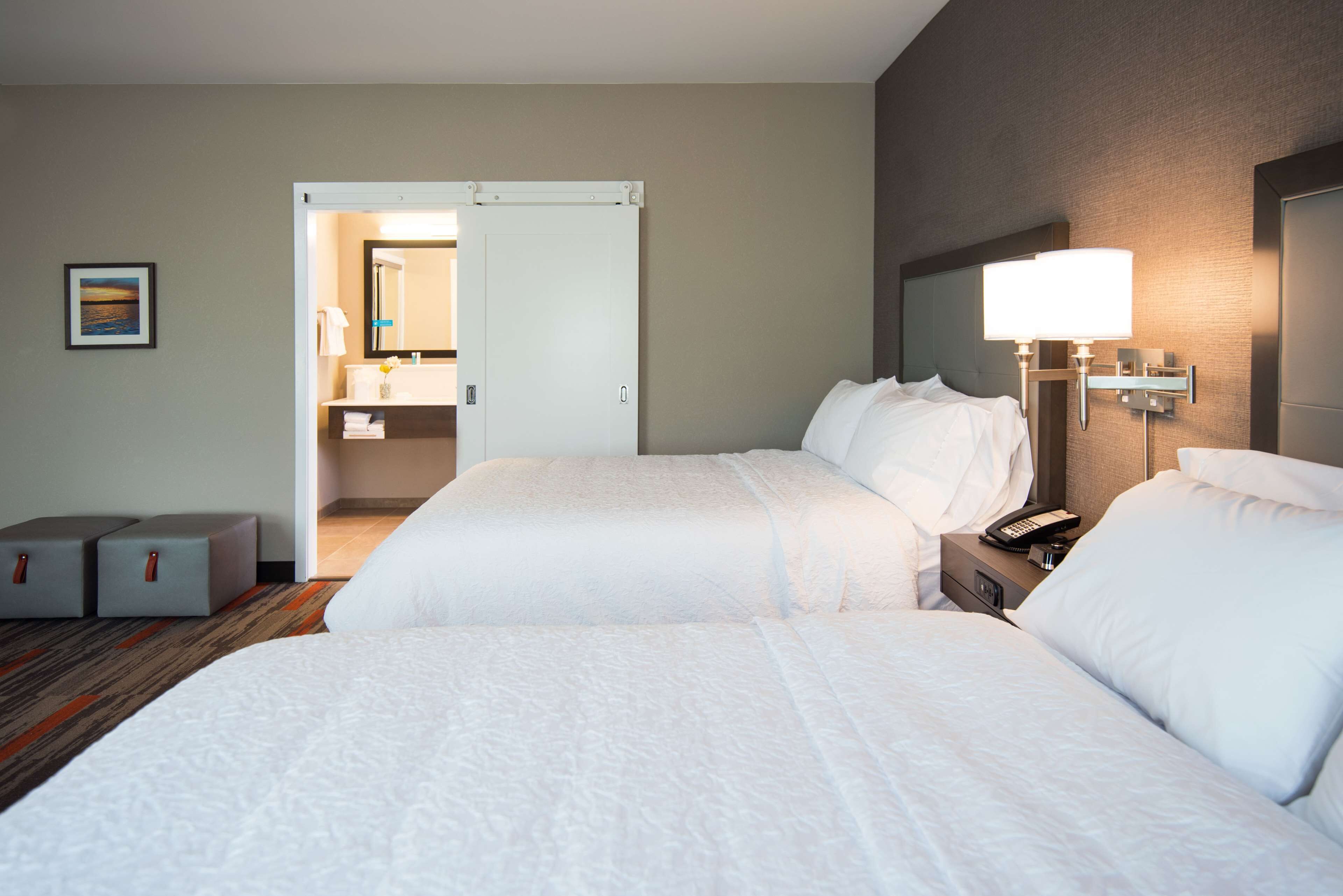 Et hotelværelse med to senge, et badeværelse, der kan ses gennem en åben dør, et sengebord med en lampe og et billede i ramme på væggen.