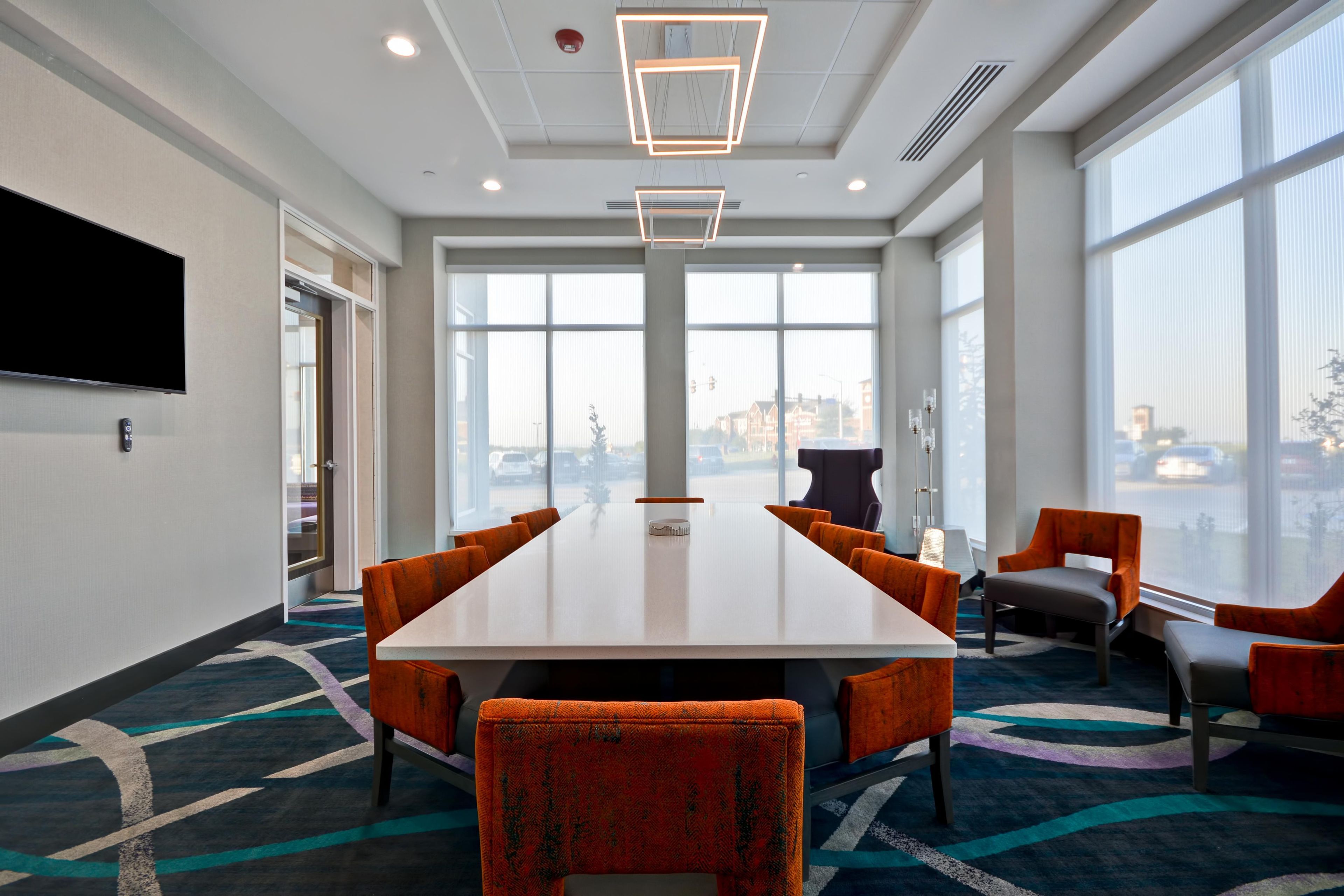 A conference room with a long table, orange chairs, large windows, and a flat-screen TV on the wall.