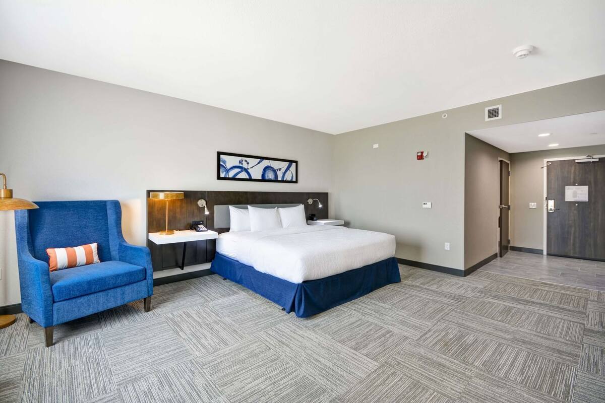 A hotel room with a bed, a desk, a chair, a painting on the wall, and a door.