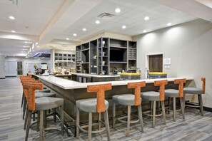 A modern bar area with a bar counter, stools, and a kitchen in the background. - Hilton Garden Inn Tulsa-Broken Arrow (Broken Arrow)