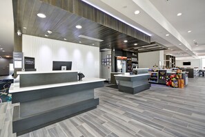 A modern hotel reception area with a grey reception desk, a digital display, and a food kiosk. - Hilton Garden Inn Tulsa-Broken Arrow (Broken Arrow)