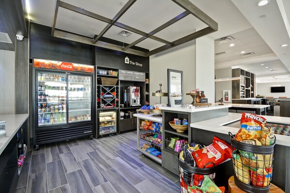 A modern retail space with a refrigerated display, snack bins, and a counter with a sign reading "The Shop". - Hilton Garden Inn Tulsa-Broken Arrow (Broken Arrow)