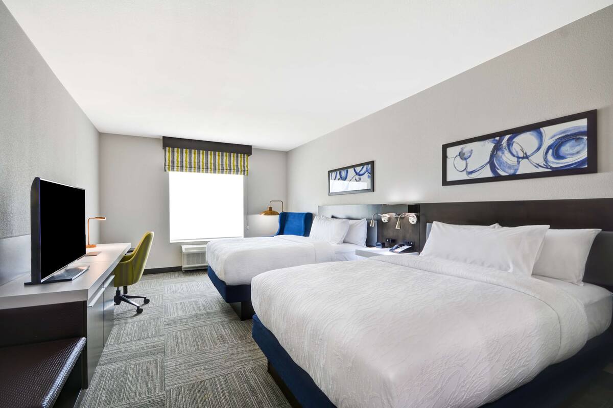 A hotel room with two beds, a desk with a computer, a TV, and artwork on the walls.