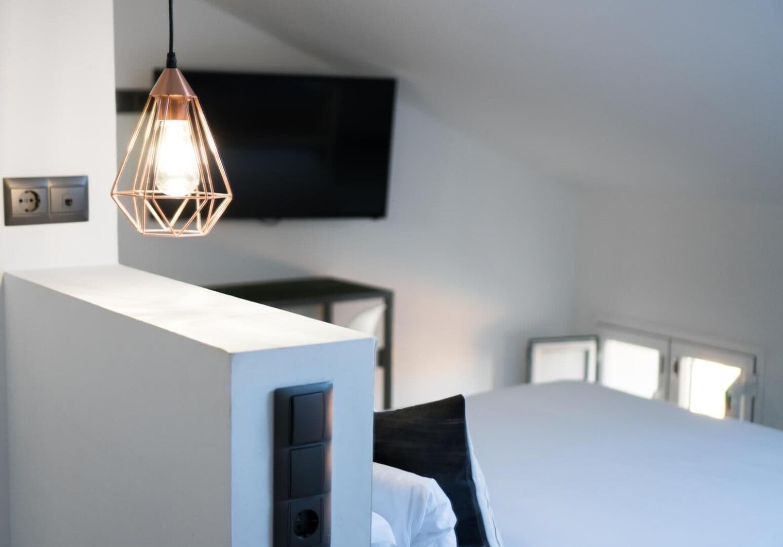 Un dormitorio moderno con cama, televisor montado y una original lámpara colgante de formas geométricas.