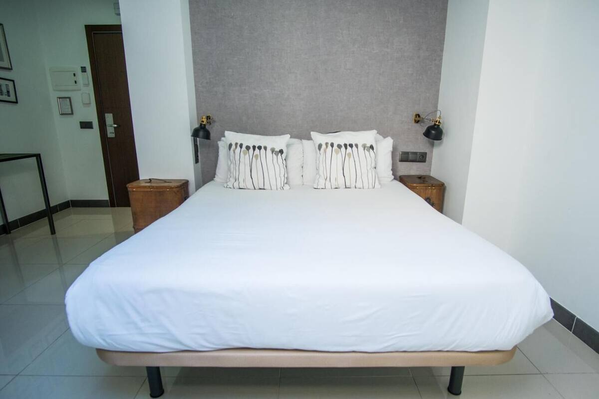 Una cama grande con ropa blanca, dos mesitas de noche con lámparas y una pared gris con textura.