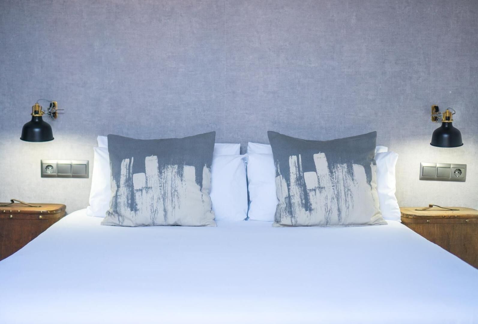 Habitación de hotel con una cama grande, dos mesitas de noche, dos lámparas de pared negras y una pared con textura.