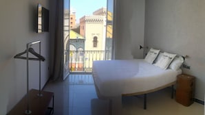 Ein Hotelzimmer mit Bett, einem kleinen Tisch, einer Hängelampe und Blick auf Gebäude durch eine Balkontür.