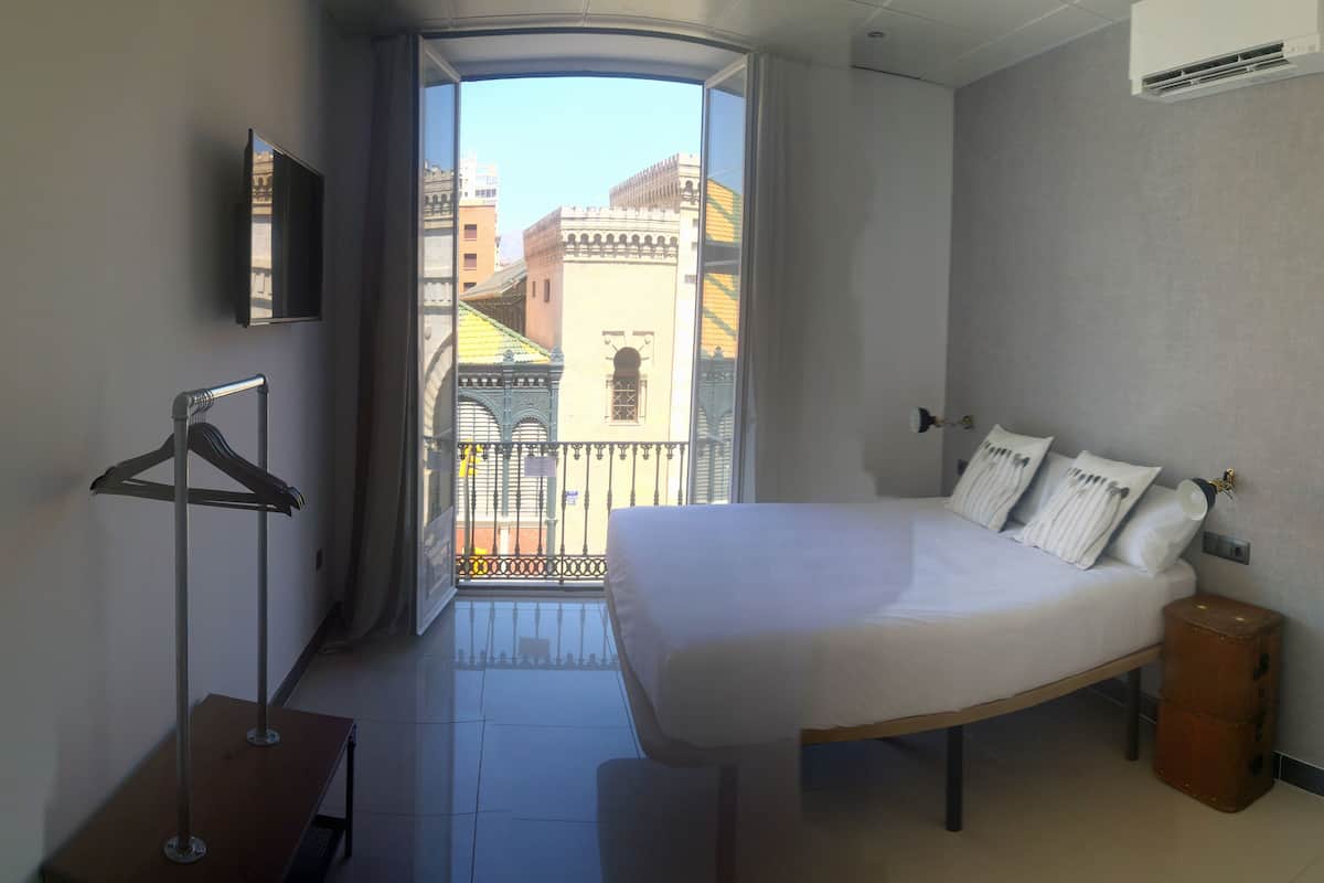 Habitación de hotel con cama, una mesita, una lámpara colgante y vistas a edificios a través de puertas de balcón.