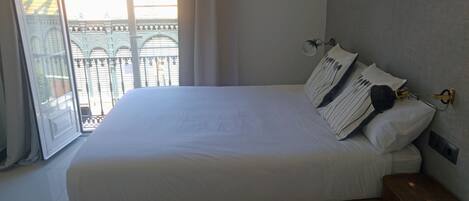 A neatly made bed with white linens and striped pillows, a wooden nightstand with a lamp, and a view of a balcony with a metal railing.