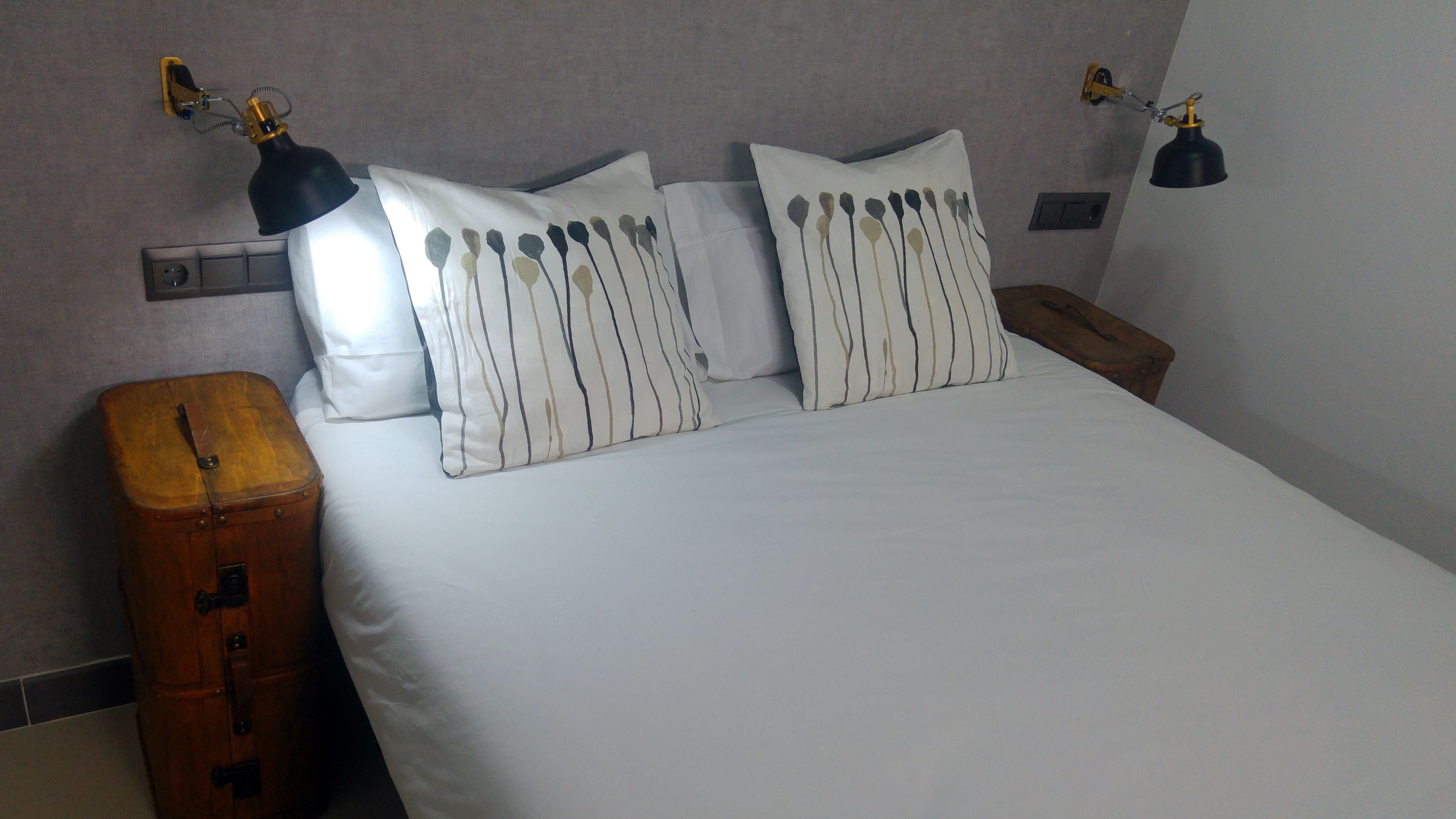 A neatly made bed with white bedding and striped pillows, two bedside lamps, and a wooden nightstand with a suitcase.