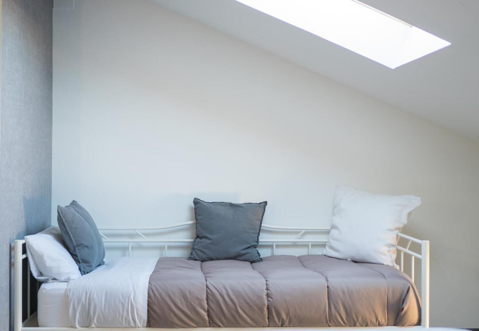 Una cama con estructura metálica blanca, colchón color beige y tres almohadas en tonos grises y blancos.