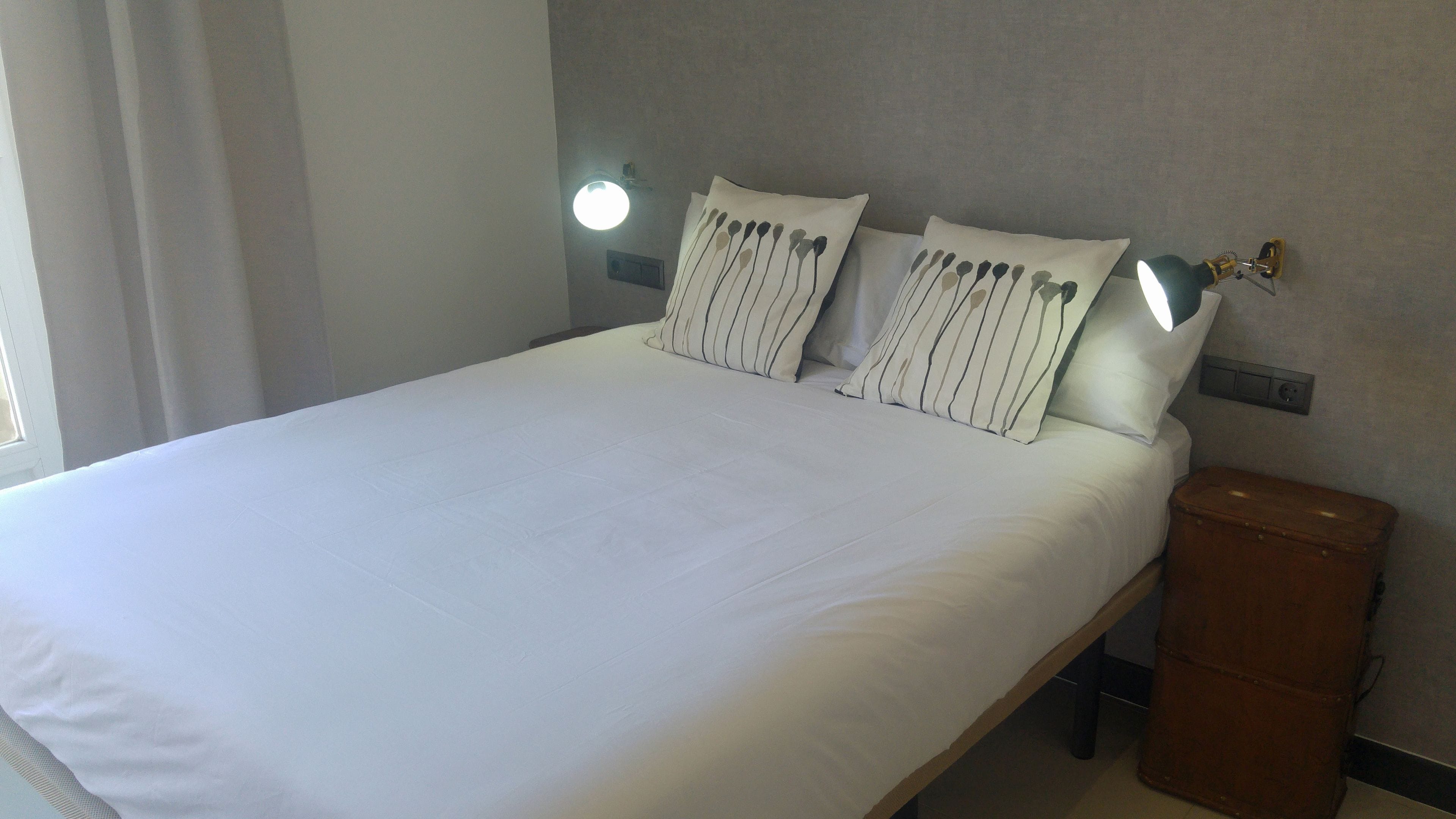 Un lit bien fait, avec du linge de lit blanc, deux oreillers à rayures, une table de chevet en bois et une valise marron.