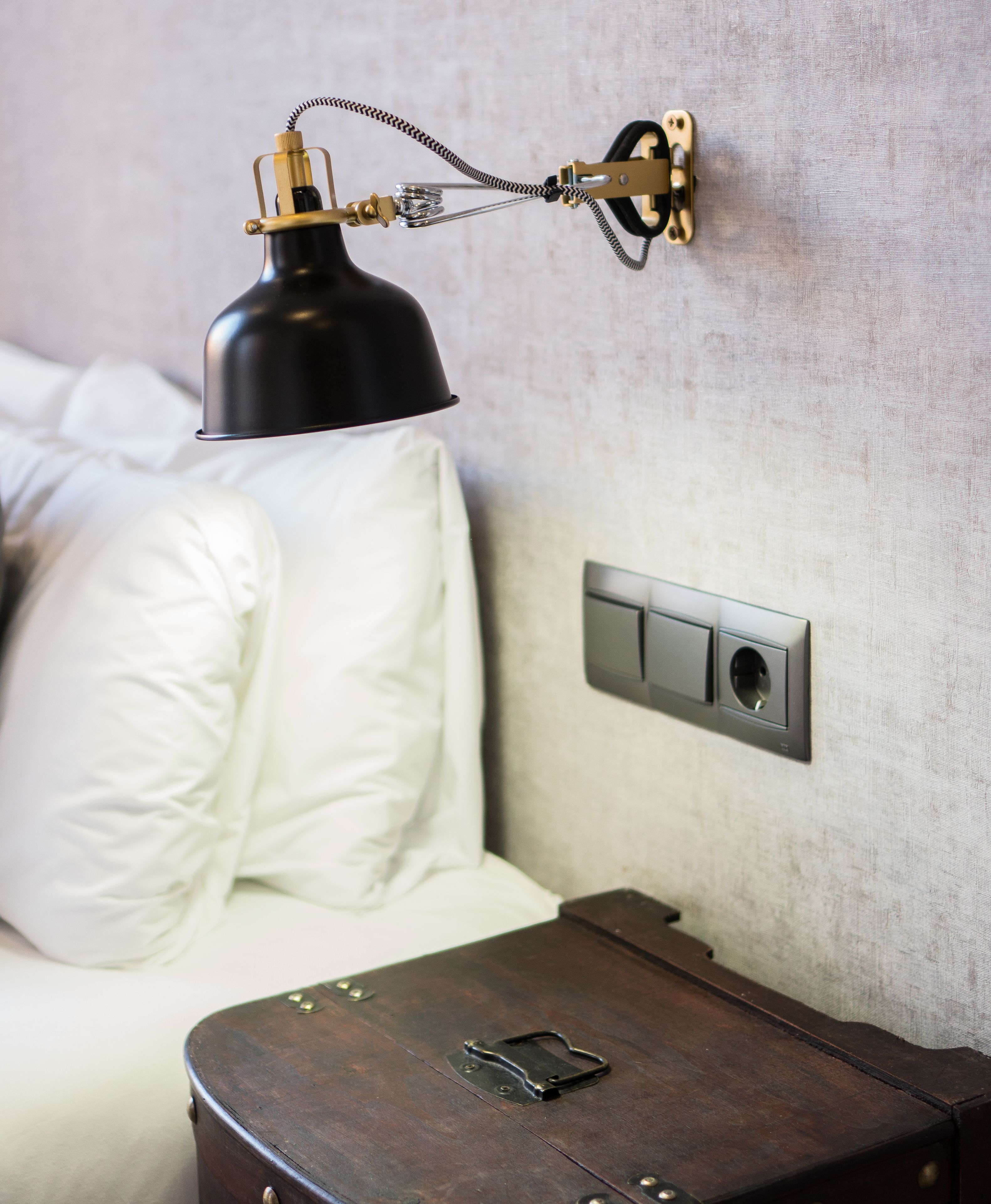 Ein Bett mit weißer Bettwäsche, eine schwarze Schreibtischlampe und ein an der Wand montiertes Schalterpaneel.