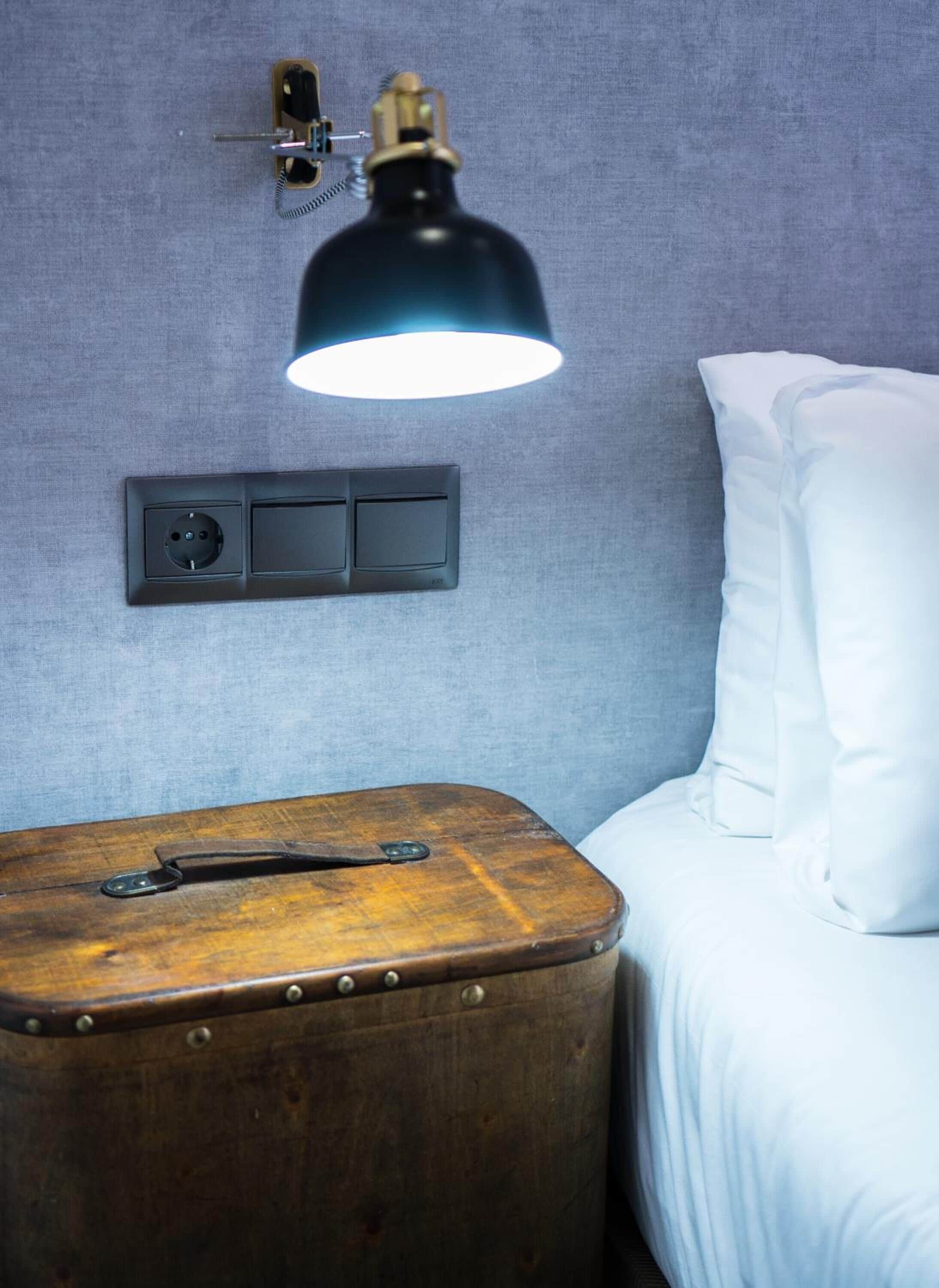 A bed with white bedding, a small wooden nightstand with a vintage lamp, and a wall socket.