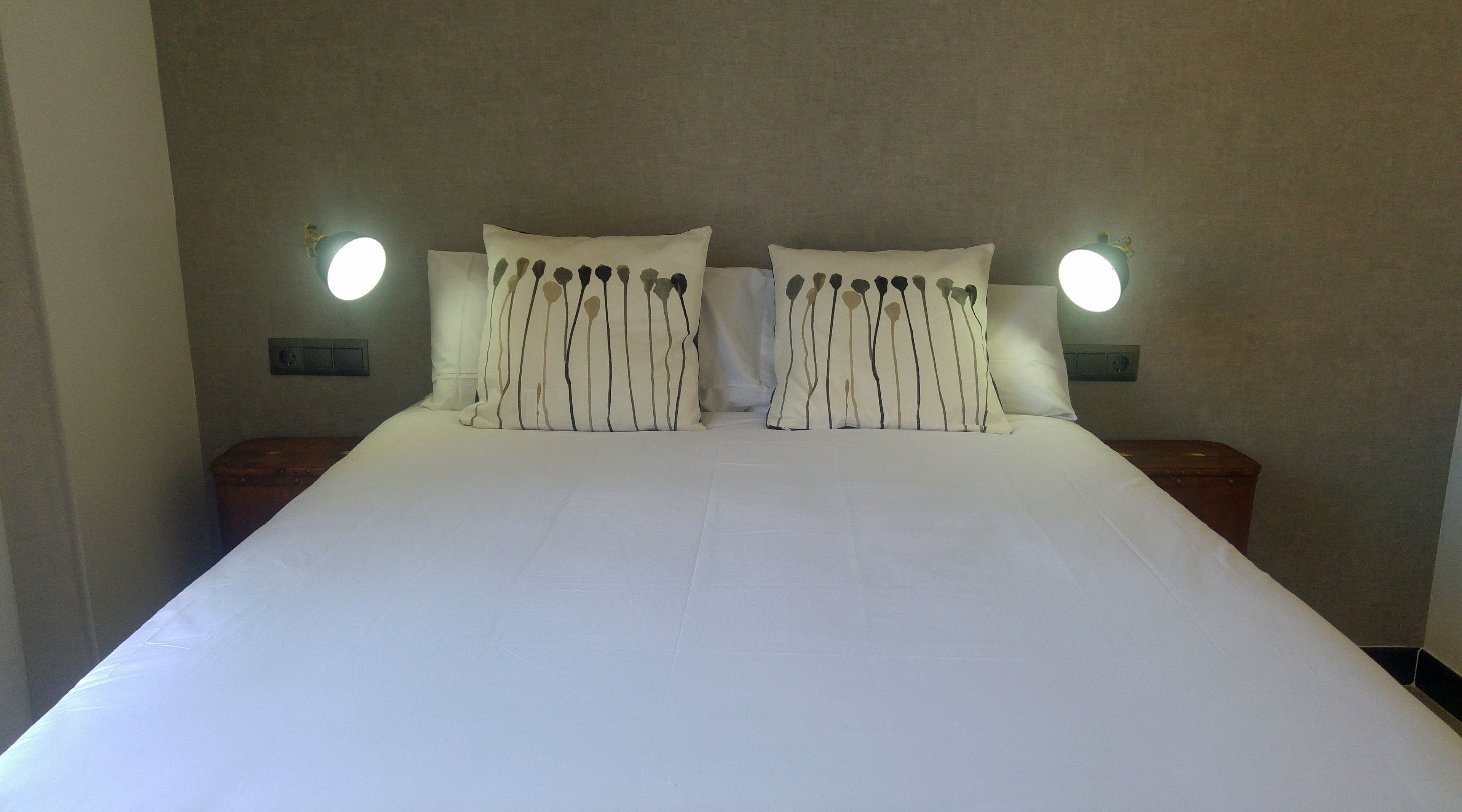 A neatly made bed with white linens and striped pillows, flanked by two bedside tables with wall-mounted lamps.