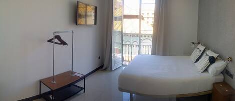 Une chambre d’hôtel avec un grand lit, un banc en bois, un porte-manteau et un balcon donnant sur des immeubles.