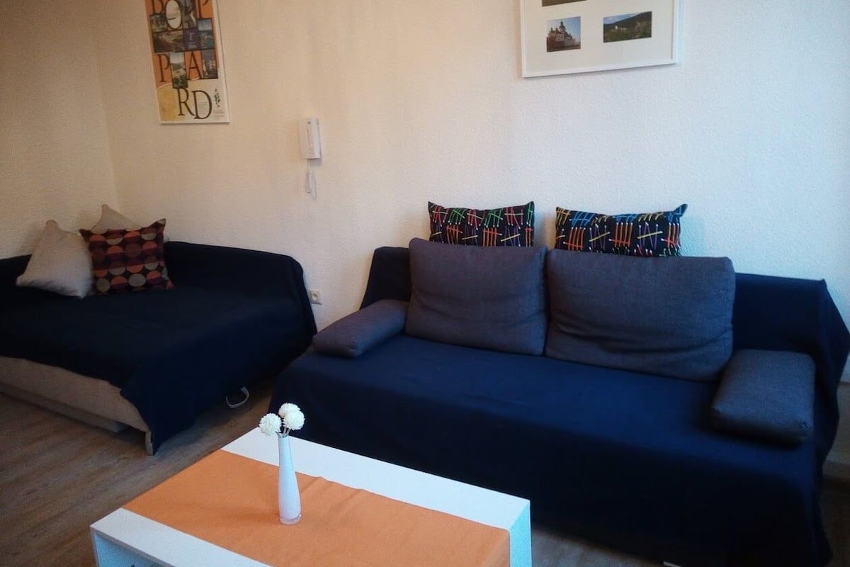 Una habitación con dos sofás oscuros, uno con cojines estampados y el otro con cojines lisos. Frente a los sofás, hay una pequeña mesa blanca con un jarrón de flores.