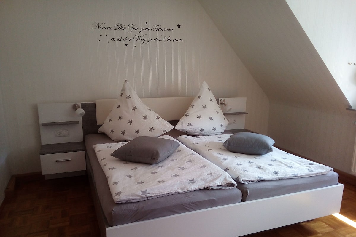 Un dormitorio con techo inclinado, una cama con cabecera blanca, dos mesitas de noche y una pared con inscripción de motivos estelares.