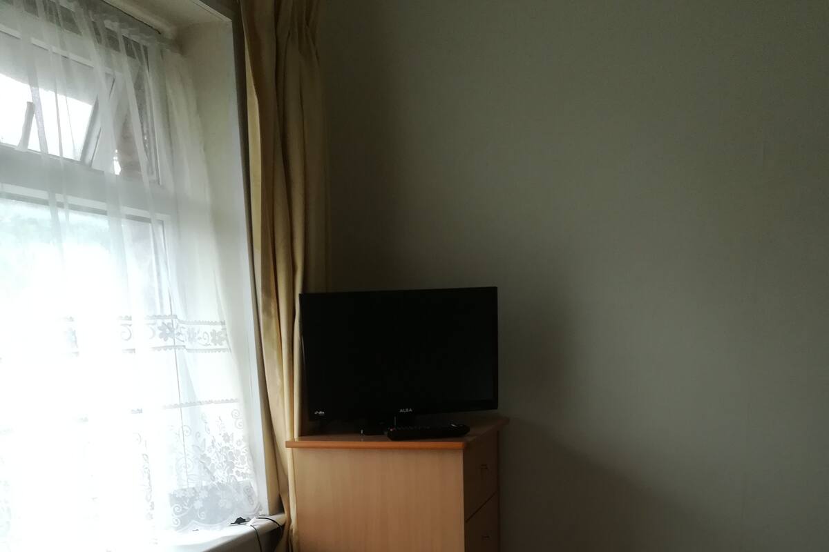 Ein Zimmer mit einem Bett, einem Fernseher auf einer Holzkommode und einem Fenster mit Vorhängen.
