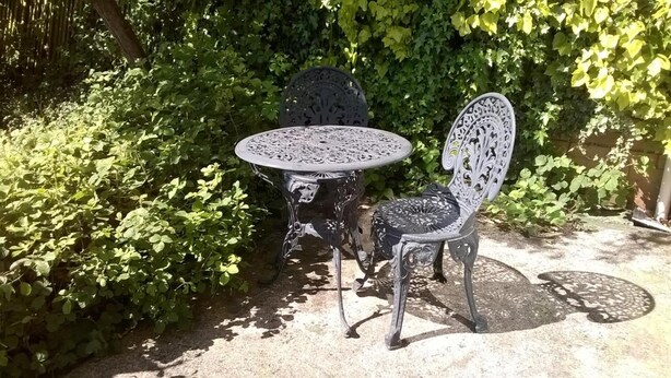 Et sæt af udendørs møbler i metal bestående af bord og stole i en have med frodig beplantning.