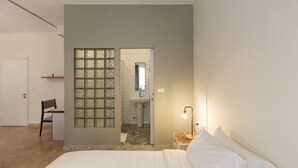 A bedroom with a bed, a chair, a nightstand with a lamp, a door, and a bathroom with a sink and mirror. - The Grand Meshmosh Hotel (Beirut)
