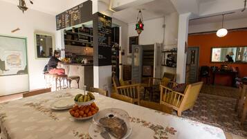 Un comedor con una mesa preparada para dos personas, un frutero y un pastel sobre un plato. Hay un espejo en la pared y una lámpara colgante.