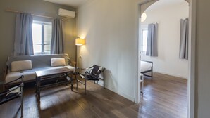A bedroom with a bed, bedside table, lamp, and a view of another room through an archway. - The Grand Meshmosh Hotel (Beirut)