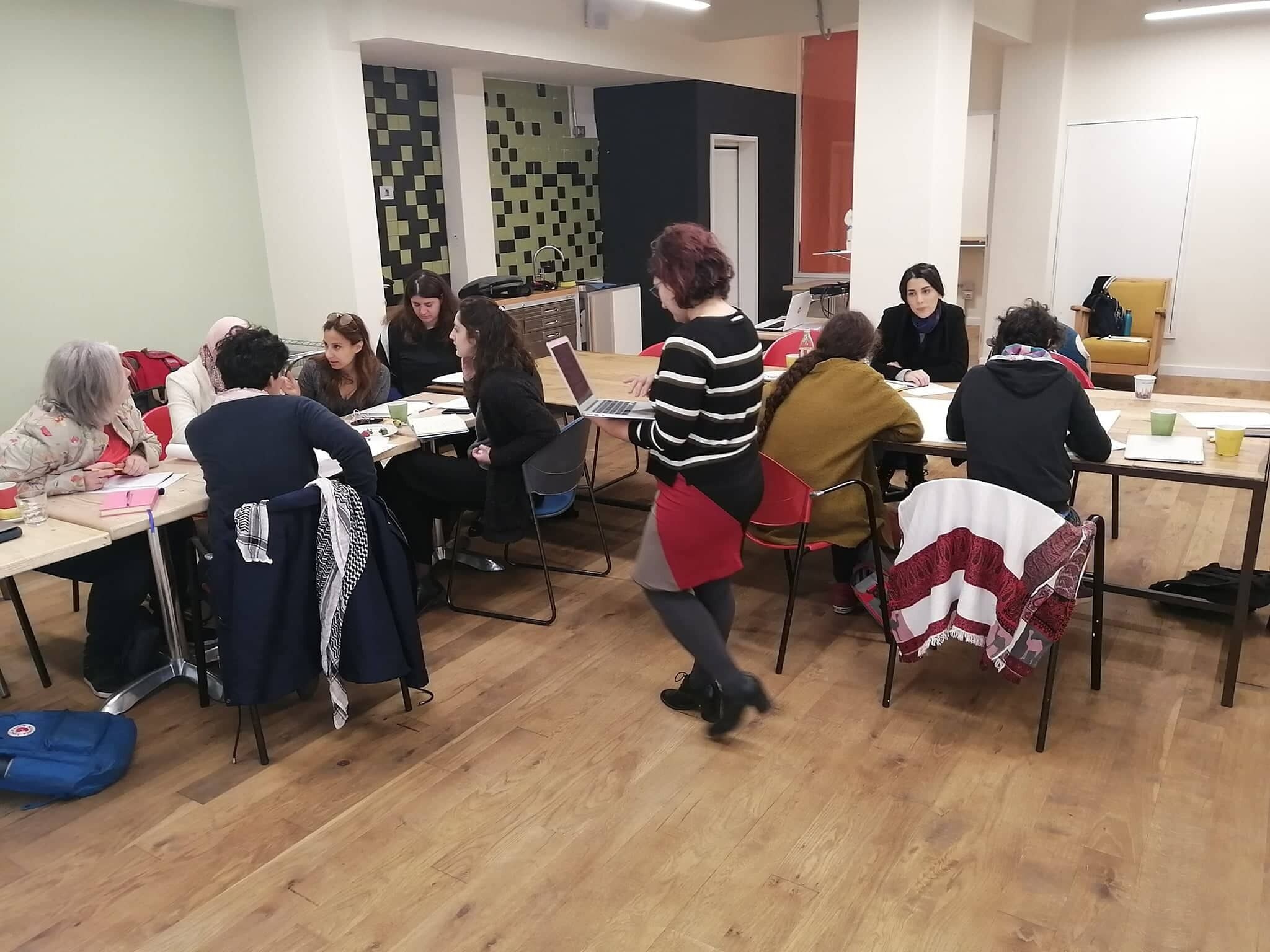 Un grupo de personas sentadas en mesas en una habitación con pared de azulejos y una puerta al fondo.