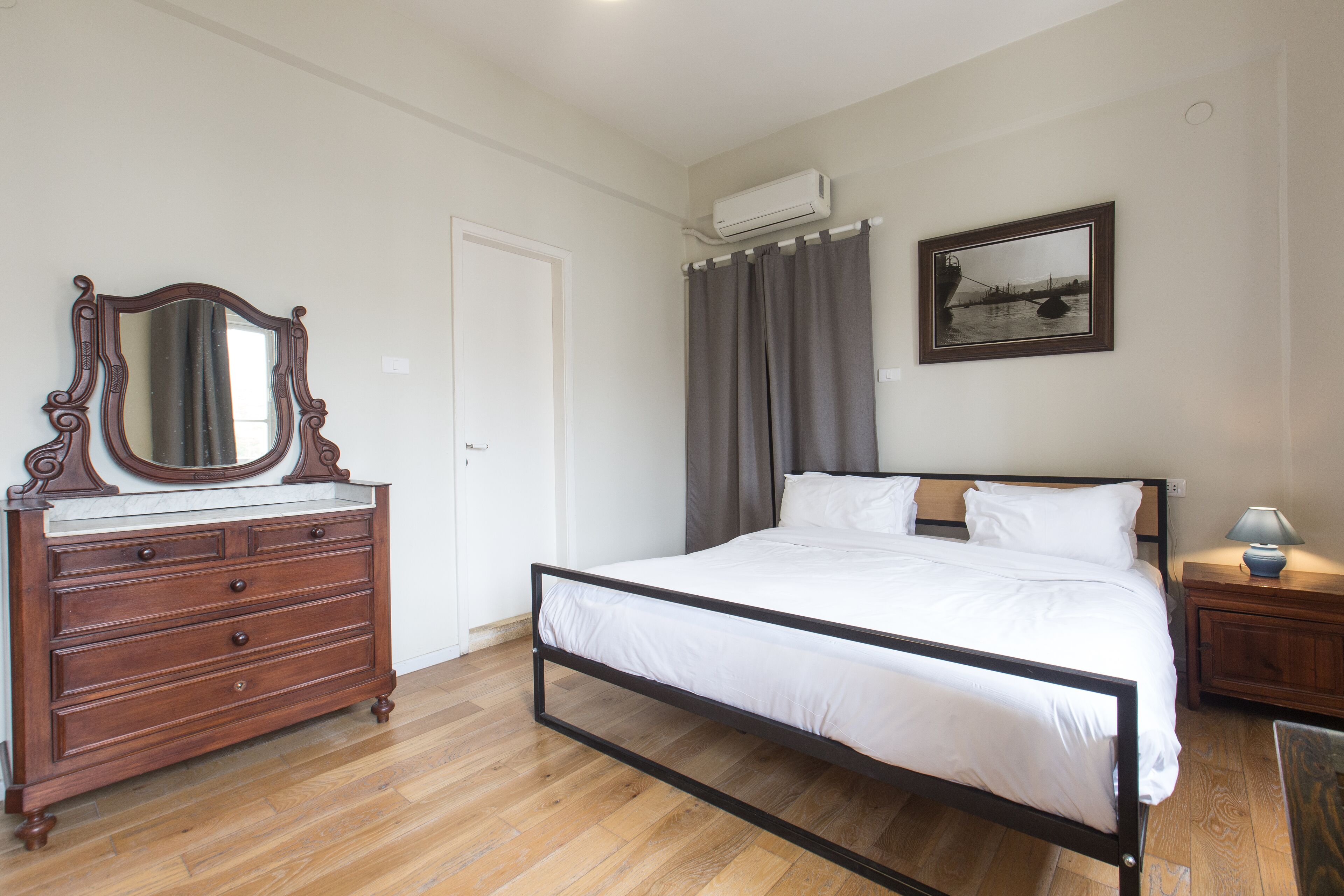 Quarto com cômoda de madeira, cama com roupa de cama branca, mesa de cabeceira, luminária, quadro na parede e aparelho de ar condicionado.