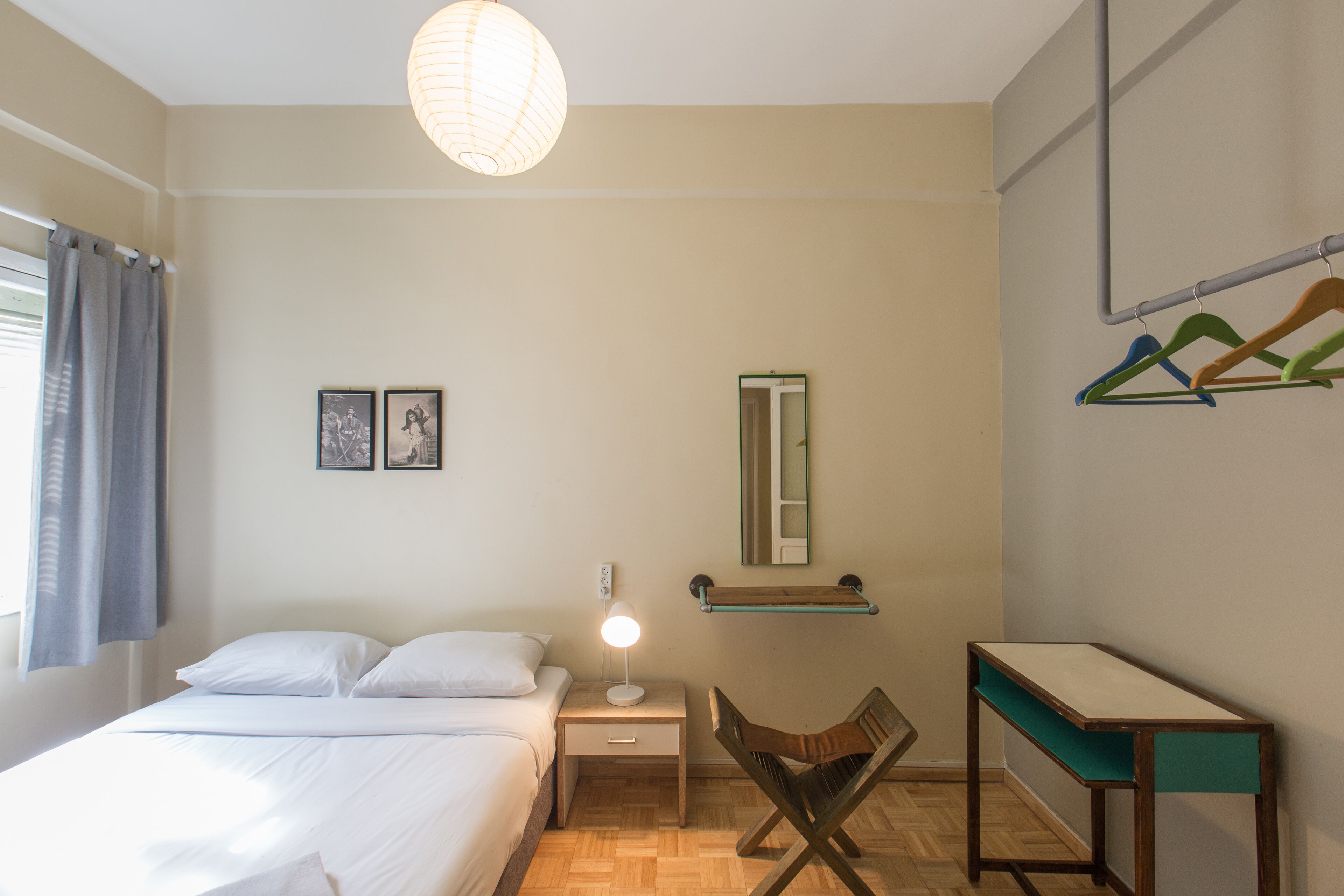 Een slaapkamer met een bed, nachtkastje, lamp, stoel, bureau, spiegel en een kledingrek aan de muur.