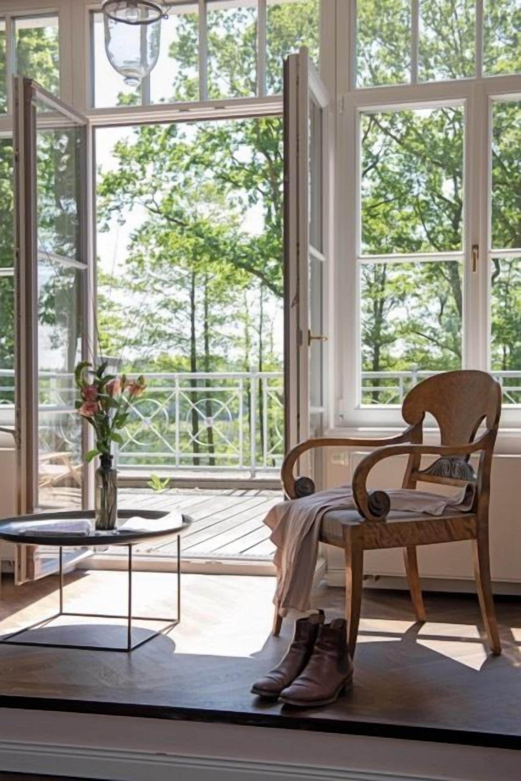 Une pièce avec une grande fenêtre, une chaise en bois, une petite table et une paire de chaussures posées sur le sol.