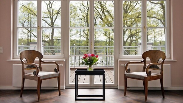 A room with two wooden chairs, a small black table, and a vase of flowers. Large windows with white frames offer a view of trees and a balcony. - Herrenhaus Röddelin (Templin)