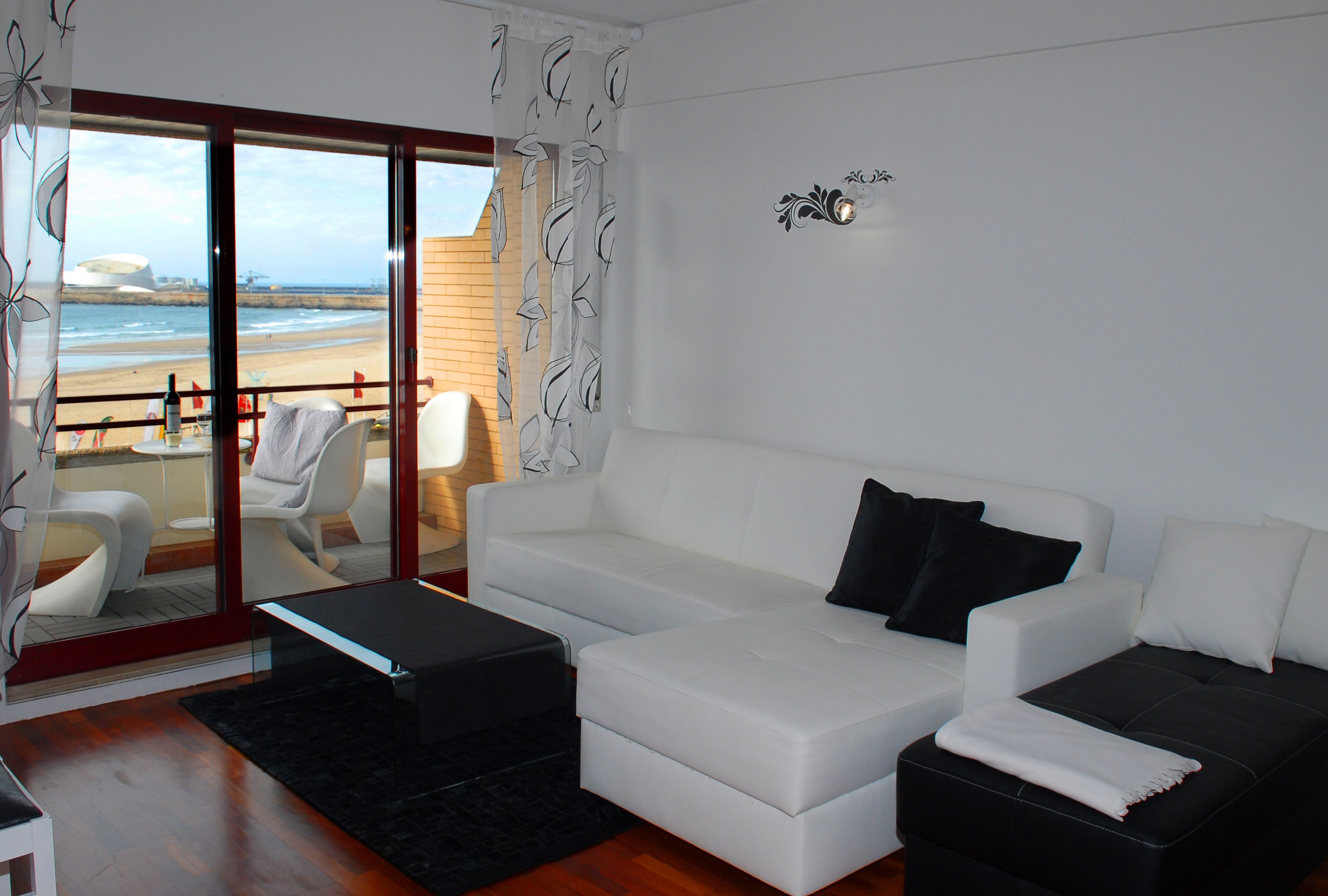 A modern living room with a white sofa, a black ottoman, and a glass door leading to a balcony with a view of the beach.