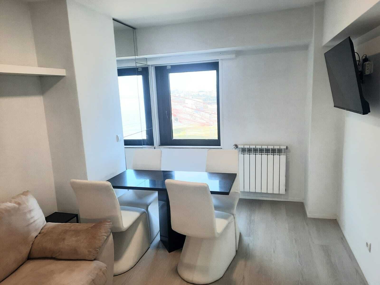 Un espace de vie moderne et compact, comprenant une table à manger et deux chaises, un canapé, une grande fenêtre et un téléviseur fixé au mur.