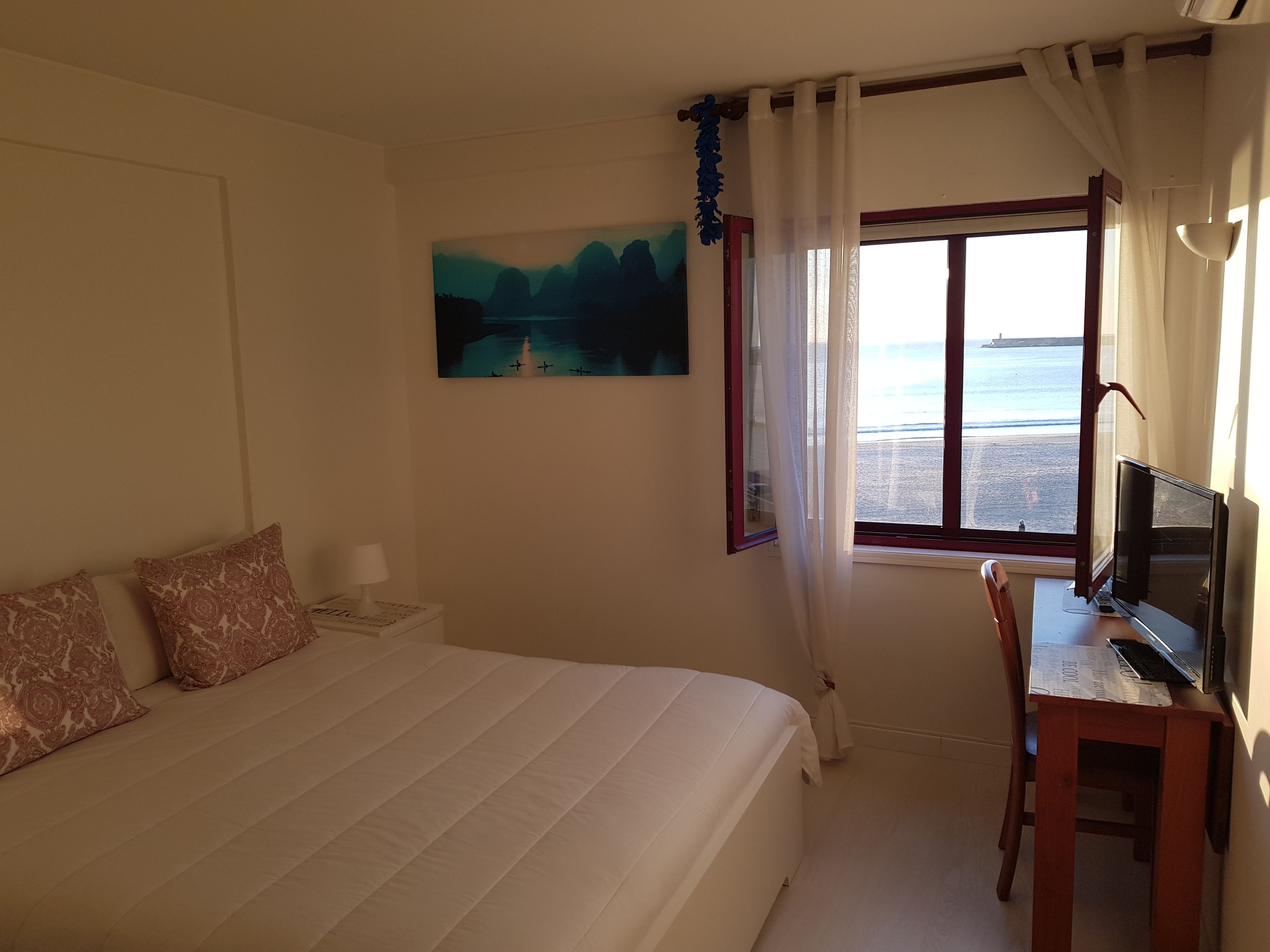 Un dormitorio con cama, un escritorio con computadora, un cuadro y una ventana con vista a la playa.