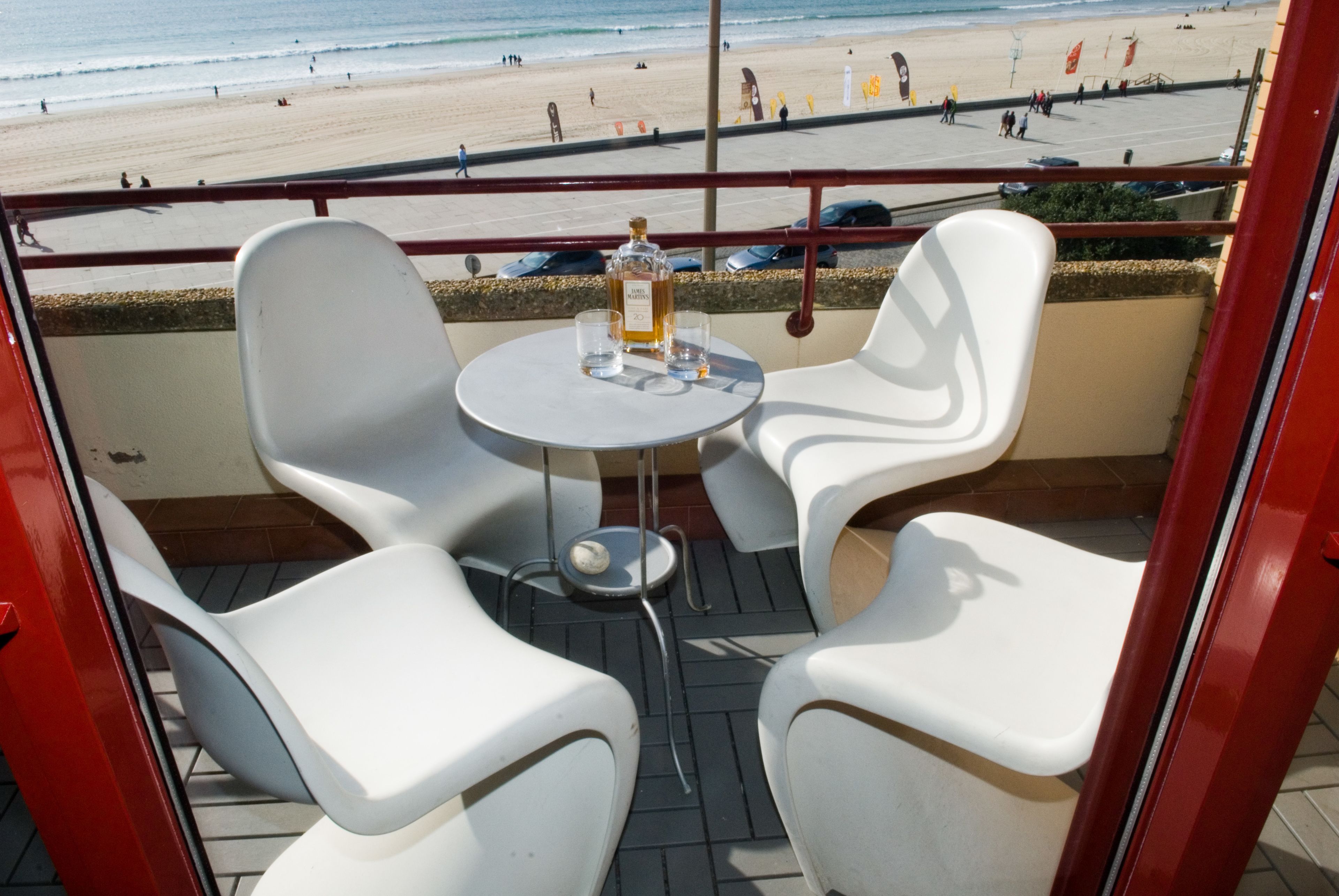 A balcony with white chairs and a table, overlooking a beach with people and umbrellas.