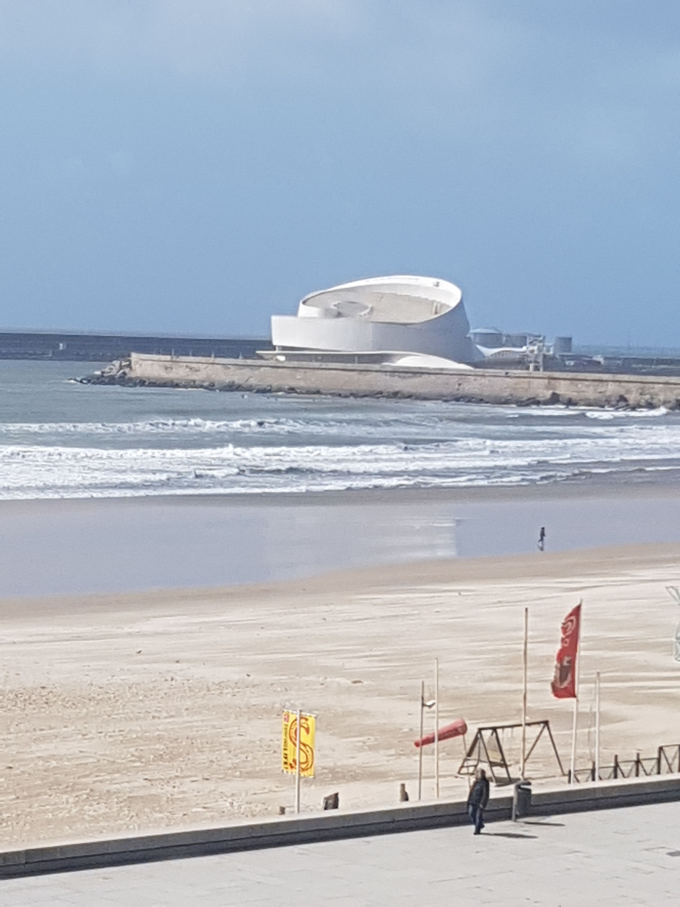 Uma praia com um edifício moderno, uma orla arenosa e algumas bandeiras.