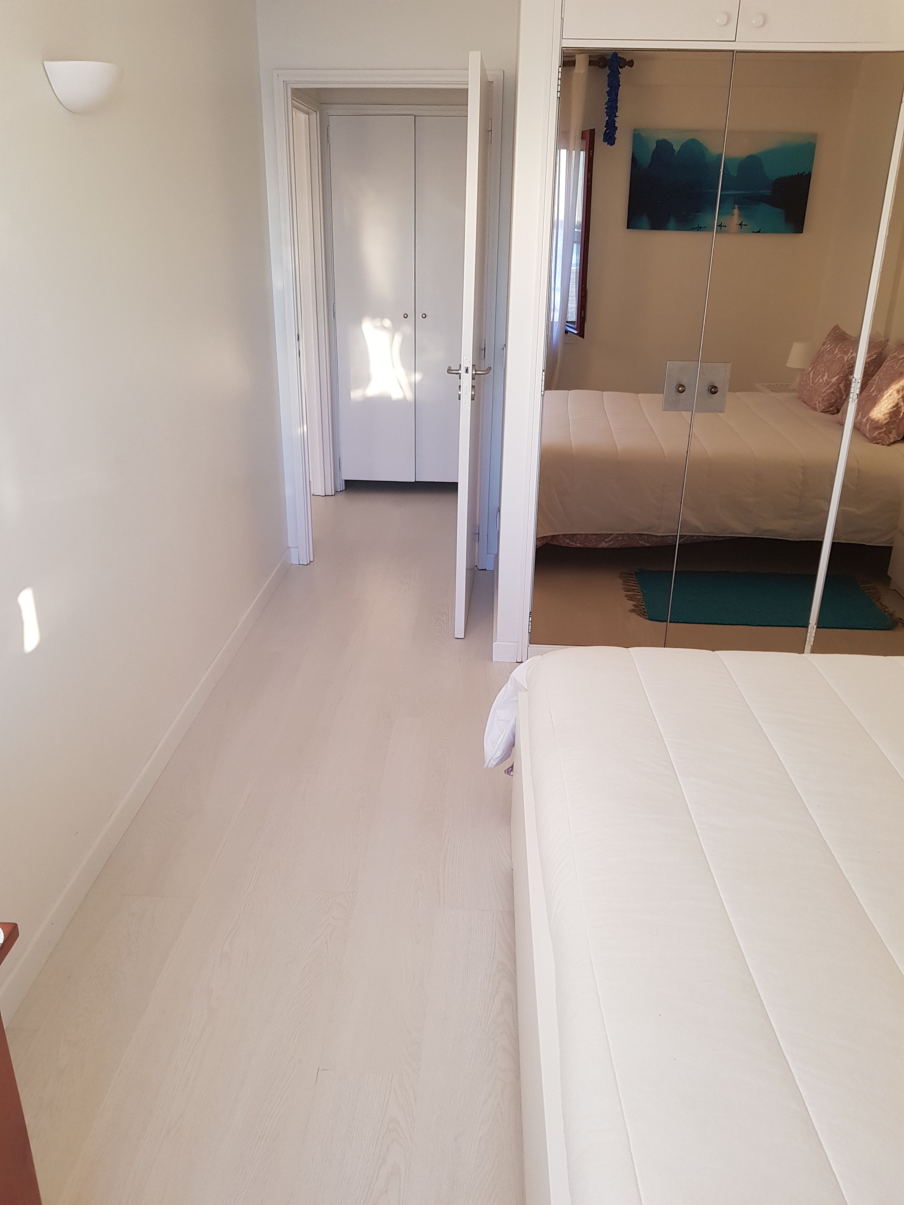 Un dormitorio con cama, mesita de noche, un cuadro en la pared y una puerta que da a otra habitación.