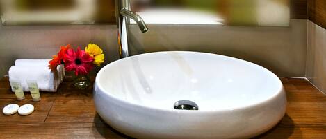 A round white sink with a modern faucet, a small vase with flowers, and toiletries on a wooden countertop.