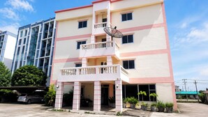 A multi-story building with a central balcony, a satellite dish, and a parking area with a car and potted plants. - UJ Apartment (Chonburi)